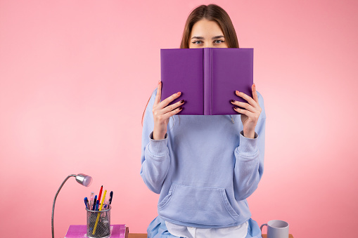 Picture of a student lovely girl with blond hair covering her face with a book of violet color and sitting on the table. High quality photo