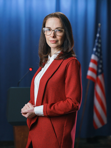 Dark haired female politician in glasses posing on camera with half smile on a blue background with American flag