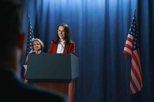Dark haired female politician during the speech on a blue background with American flags