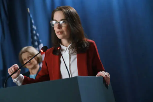 Photo of Female American politician in a red jacket at the debates, seriously responding the question from the audience