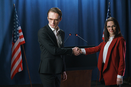Young white American politicians at the debate, shaking hands, we see them against the blue background with American flags