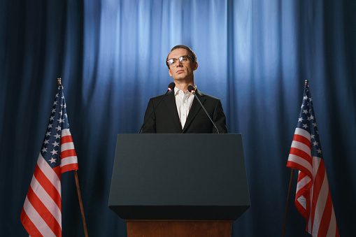 Low angle view at the serious young American politician looking away during his speech at the debates, standing behind the pedestal on a blue background with two American flags