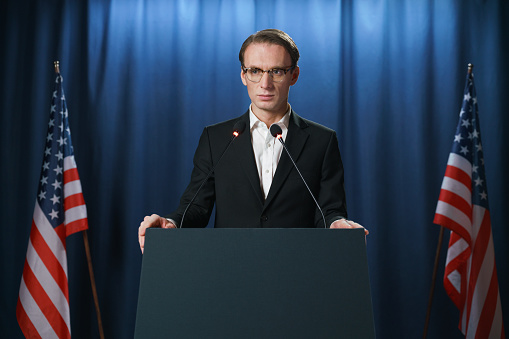 Serious young American politician looking away during his speech at the debates, standing behind the pedestal on a blue background