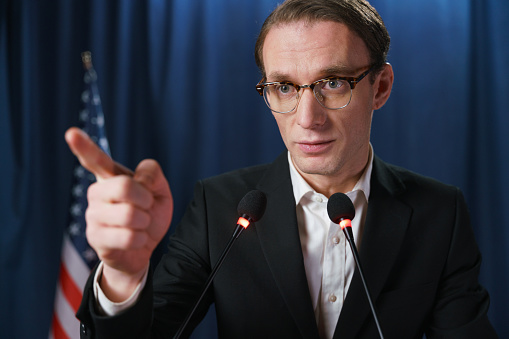 Close-up of a young emotional politician pointing by his index finger during the speech at the debates on a blue background