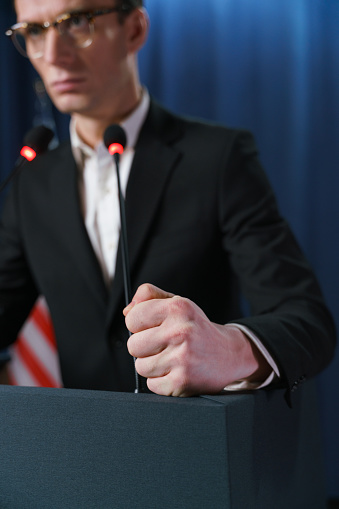 Close-up of a young politician's fist on pedestal during the speech, we see a man as a blurred background, only his fist is in focus in the foreground
