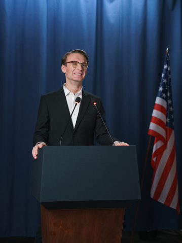 Young white politician smiles during his speech at the debates, we see him on a blue background with American flag