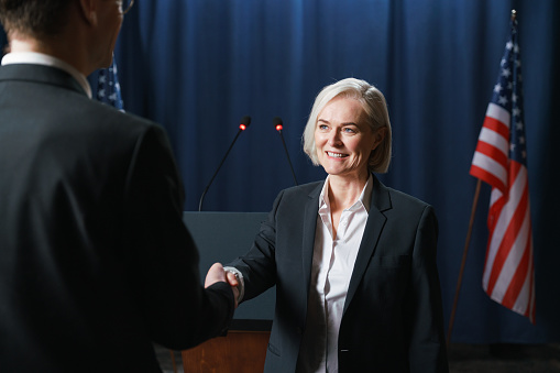 Confident blond female delegate in eyeglasses and grey suit looking at audience during speech while sitting against foreign colleagues