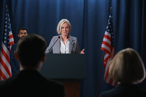 blond female politician giving a speech at the debates, standing on a stage with blue background - press conference public speaker politician speech imagens e fotografias de stock
