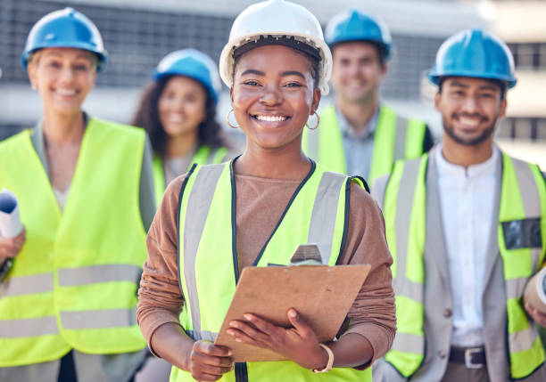 retrato recortado de una atractiva trabajadora de la construcción de pie en un sitio de construcción con sus colegas en el fondo - industrial laborer fotografías e imágenes de stock