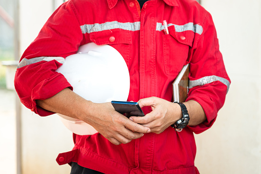 An engineer or manager in red uniform is holding smartphone and white safety helmet during take group meeting with team staff. Industrial working people in action photo. Selective focus at hand part.
