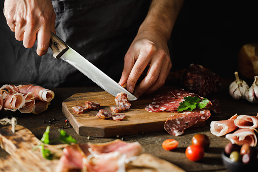 Close-up of a woman hands cuts salami sausage on wooden board.  Female hands preparing cheese and meat platter on wooden kitchen table.