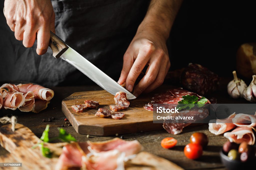 Close-up of a woman preparing cheese and meat platter Close-up of a woman hands cuts salami sausage on wooden board.  Female hands preparing cheese and meat platter on wooden kitchen table. Meat Stock Photo