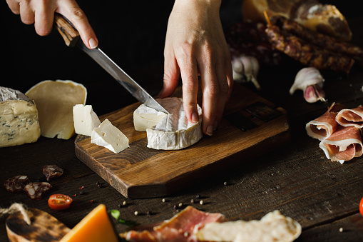 Woman hand cutting camembert on wooden board with other ingredients on table.