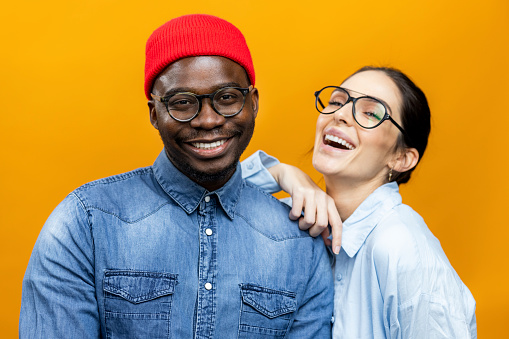 Portrait of an African American male fashion model and a Caucasian female fashion model posing for a photo shoot in a professional studio with an orange background, smiling