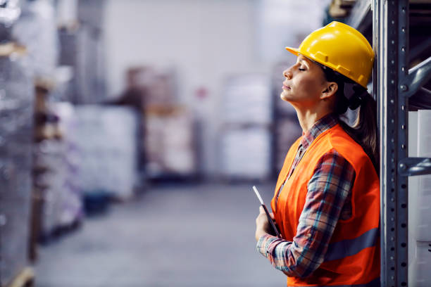 Tired female worker with tablet in hands leaning on shelf and taking a break in warehouse. stock photo
