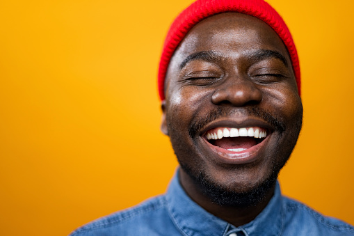 Close up portrait of a young African American male photo model posing for a photo shoot in a professional model studio with an orange background, wearing denim and a red knit hat and smiling with his eyes closed