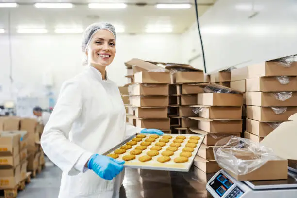 Happy female food plant worker holding tray with baking biscuits and smiling at the camera.