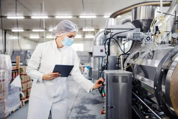 A female milk plant operator holding tablet and adjusting milk processing machine during corona virus.