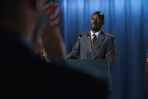 Young African-American politician in a gray suit with a speech at the debates, standing behind the pedestal against the blue background, we see him from the waist up from the distance