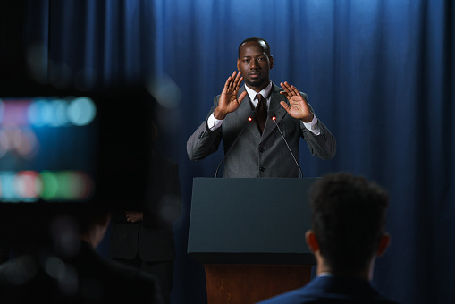 Young serious African-American speaker in a gray suit gesturing during his speech at the debates with a shooting camera in the foreground, he is standing behind the pedestal against the blue background, we see him from the waist up from the distance