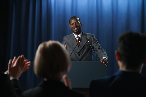 Young African-American politician in a gray suit smiles to the audience after his speech at the debates, he is standing behind the pedestal against the blue background, we see him from the waist up from the distance