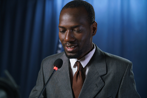Close up of a smiling dark-skinned politician in a gray suit during the speech, standing against the blue background with American flag