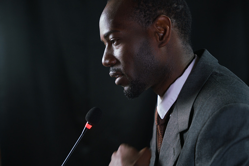 Close-up profile of a young dark-skinned politician in a gray suit speaking in microphone at the debates