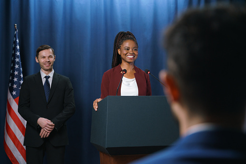 Young dark-skinned female politician in a red suit giving a speech with a smile, standing at the stage with a blue background, we see her from the distance