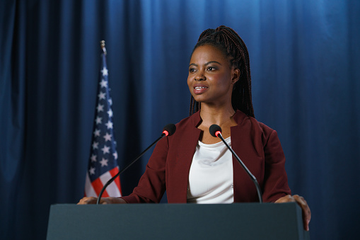 Young dark-skinned speaker during her performance at the debates, standing against the blue background with American flag