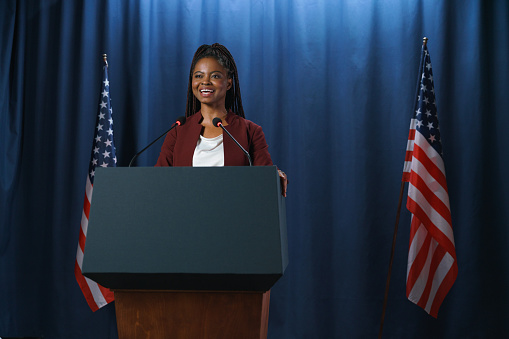 Smiling African - American student in a red suit at the debates, standing at the stage with two American flags from both sides