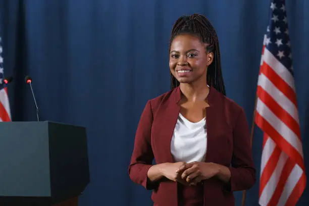 Beautiful African politician in a red suit posing on camera with a smile before  the speech, standing against the blue background with American flags