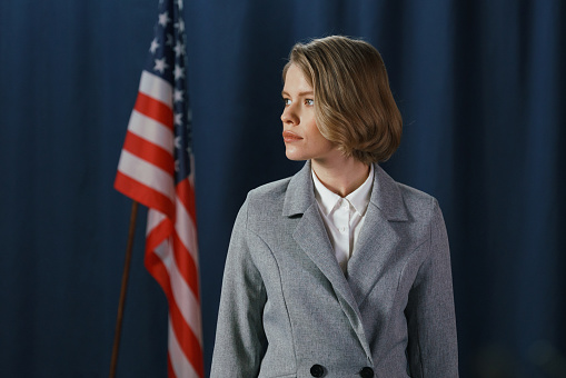 Serious white female politician in a gray suit looking away, standing on a blue background with American flag