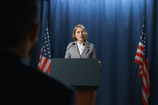 Young blond female politician gives a speech standing on a stage behind a pedestal on a blue background with American flags, we see her from the waist up