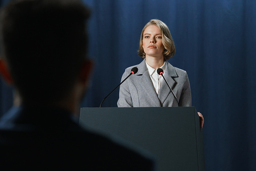 A young American female politician during the speech at the debates standing on a stage behind a pedestal on a blue background and a listener's shoulder in the foreground