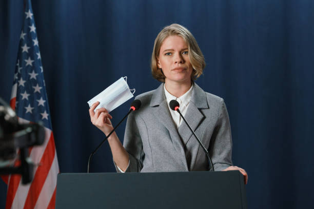 istock Young American female speaker with a protective mask in hand during her speech on a dark blue background 1377435144