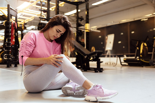 Shot of a sportswoman with a knee injury. Front view of athletic woman holding her knee in pain on sports training in a health club. Cropped shot of a woman suffering from a knee injury.