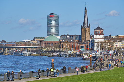 Düsseldorf, Germany - February 23, 2022: People at the Rhine promenade in Dusseldorf on a sunny winter day. In the background the Tower Museum and the Church of St. Lambertus.