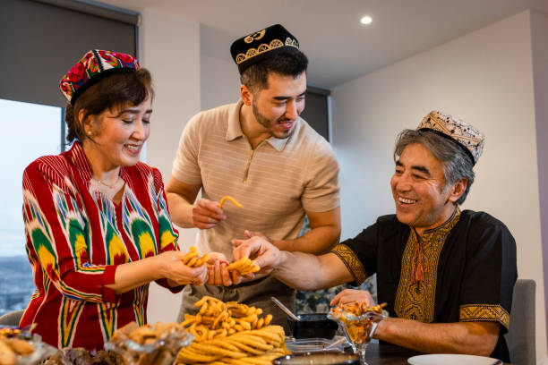 Sharing the Sangza A senior Uyghur couple and their son wearing traditional clothing and Doppa's, a traditional hat of the Uyghurs. They are sharing and eating sangza, deep-fried noodles in a twisted pyramid shape, at the dining table to celebrate Eid al-Fitr the end of Ramadan. central asian ethnicity stock pictures, royalty-free photos & images