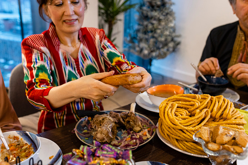 A medium shot of a senior Uyghur woman wearing traditional clothing and a Doppa, a traditional hat of the Uyghurs. She is breaking a naan bread to share with her family at the dining table to celebrate Eid al-Fitr the end of Ramadan.