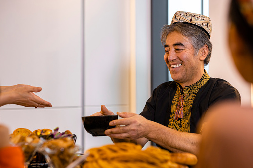 A medium shot of a senior Uyghur man wearing traditional clothing and a Doppa, a traditional hat of the Uyghurs. He is passing one of his family members a bowl of dumpling soup at the dining table while they celebrate Eid al-Fitr the end of Ramadan.