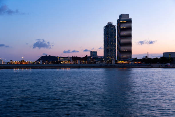 Illuminated buildings along Barcelona's Port Vell harbor at night, reflecting city lights on the calm water, with the iconic Columbus Monument towering in the background