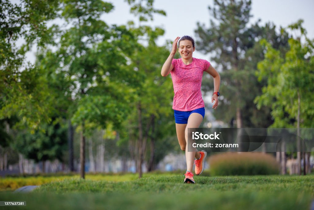 Happy young woman in running shorts jogging on grass padded track in public park Happy young woman wearing running outfit jogging on grass padded track in public park in the city, life in motion 18-19 Years Stock Photo