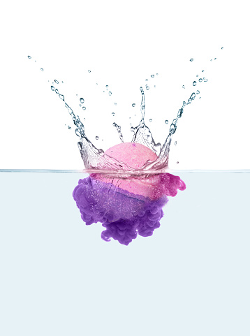 Composite image: a pink and purple bath bomb half in and half out of water with water splash above and dissolve effect below.