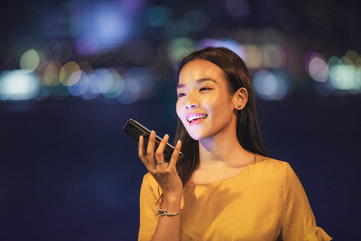 A woman smiling as she records a voice message to a friend, from the Hong Kong harbourfront at night.