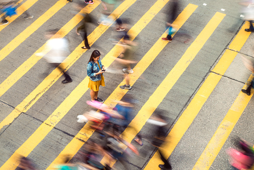 A woman pausing to focus on a message on her phone while on a busy crosswalk, with motion blur as people move around her.