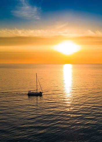 Yacht sails to the horizon in the setting sun - photograph of a bright sunset with a yacht sailing on the sea. Beautiful sunset at sea over the horizon with clouds in the sky. A small boat sails into the sunset. A sunset over the ocean. Reflection of a sunset at sea. Sunrise at sea. Sunset in the clouds over the sea horizon. Little white boat floating on the water towards the horizon in the rays of the setting sun. Beautiful clouds with yellow highlights.