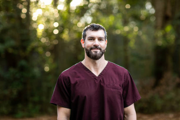 A doctor with dark hair and a beard in maroon scrubs standing outside in a natural green environment A doctor with dark hair and a beard in maroon scrubs standing outside in a natural green environment. red medical scrubs stock pictures, royalty-free photos & images
