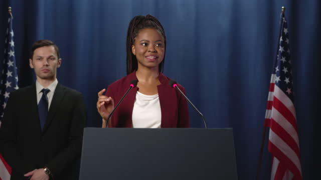 Young African American politician in a red jacket thanks the audience after a speech while camera zooms her in