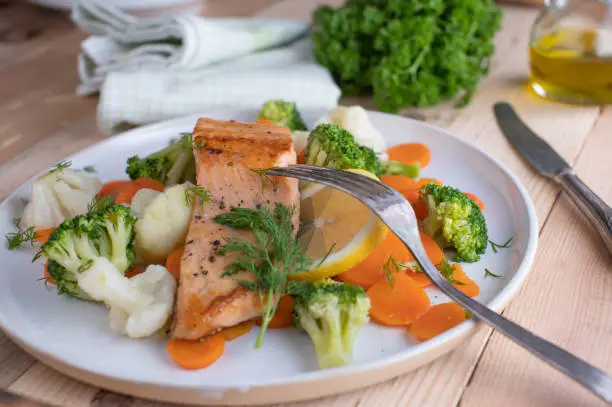 Homemade fresh cooked healthy salmon dish with imperial vegetables such as broccoli, cauliflower and carrots. Served on a white plate on wooden kitchen table. Low carb meal for dieting