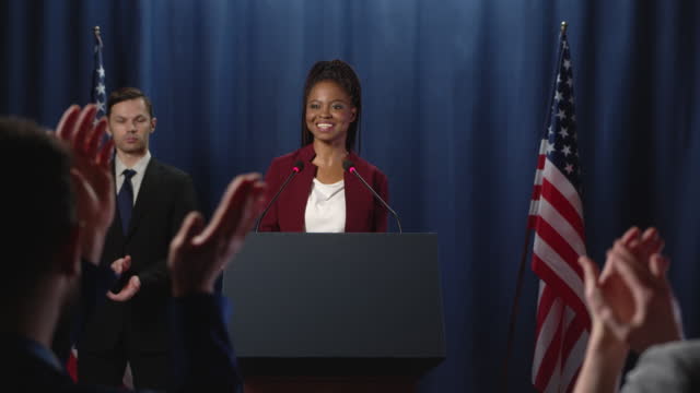 A young female African American politician in a red jacket thanking the audience for applause after a speech